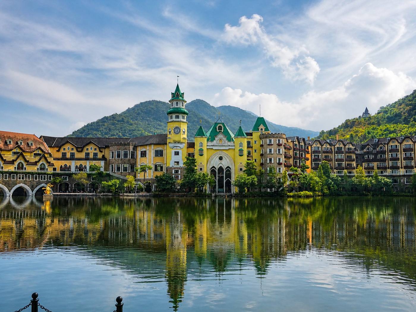 The exterior of The Interlaken OCT Hotel Shenzhen across the Tea Stream Valley lake, yellow Bavarian alpine facades, green copper-roofed clock tower and turrets reproducing Interlaken, Switzerland, half-timbered townhouses, a golden dragon sculpture on the lakefront, and the Yantian District forested mountains rising behind the resort